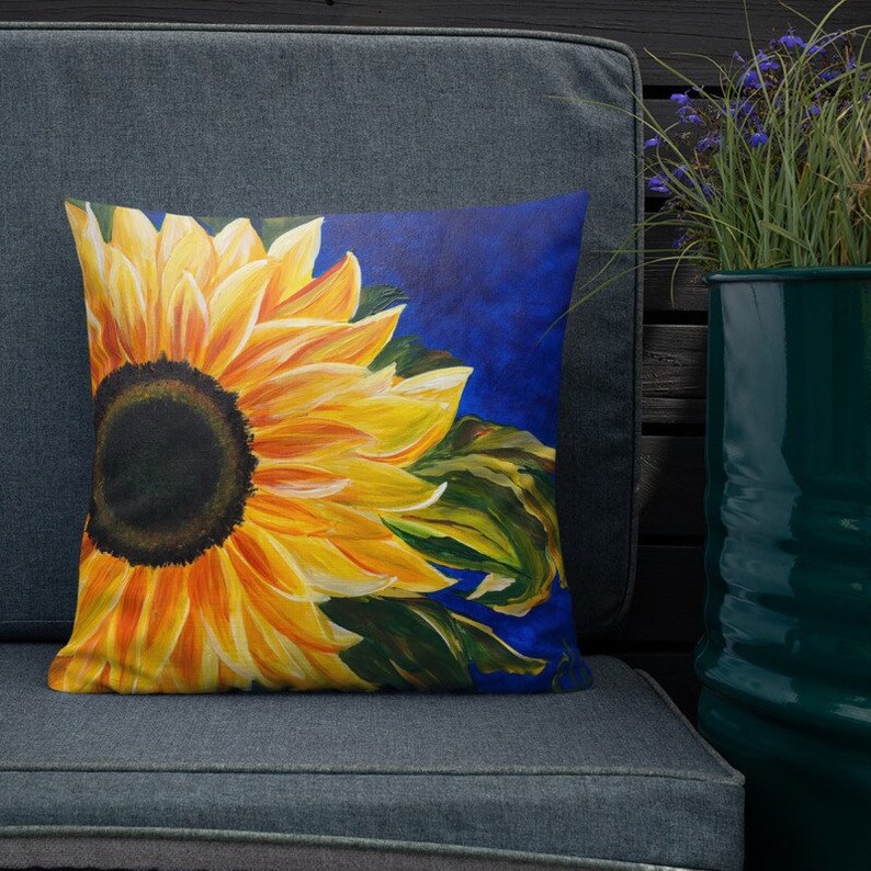 May include: A throw pillow with a vibrant yellow sunflower on a blue background. The sunflower is painted in a realistic style with a dark brown center and yellow petals.