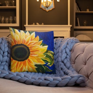May include: A throw pillow with a vibrant sunflower design. The sunflower is painted in shades of yellow and orange, with a dark brown center. The background is a deep blue.