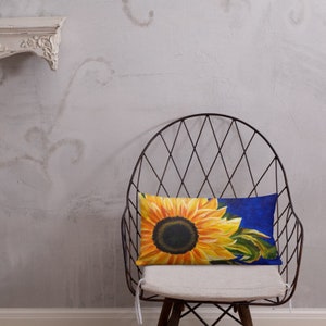 May include: A throw pillow with a sunflower design on a blue background. The sunflower is yellow with a dark brown center and green leaves. The pillow is sitting on a chair with a wire frame.