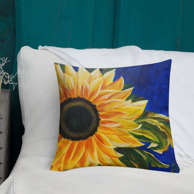 May include: A throw pillow with a painting of a sunflower with yellow petals and a dark brown center. The sunflower is on a blue background with green leaves.