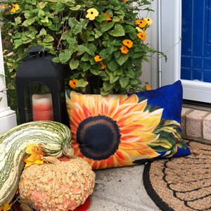 May include: A decorative doormat with a sunflower design, a black lantern with a candle, a gourd, and a striped squash. The doormat is made of natural fibers and has a black border. The lantern is made of metal and has a glass panel. The gourd is orange and has a bumpy texture. The squash is green and white and has a striped pattern.