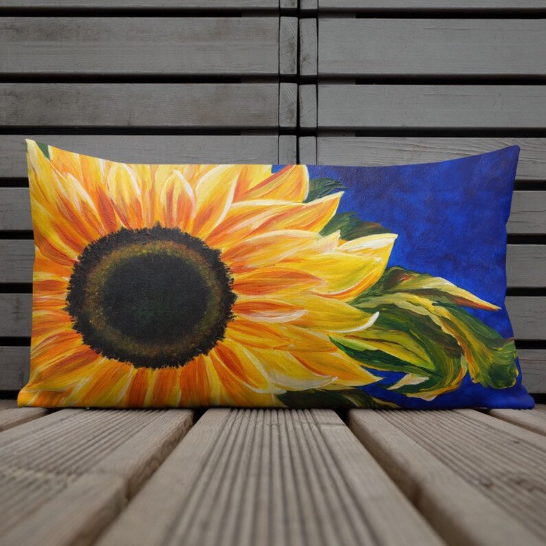 May include: A rectangular pillow with a vibrant sunflower design. The sunflower is painted in shades of yellow and orange, with a dark brown center. The background is a deep blue color.