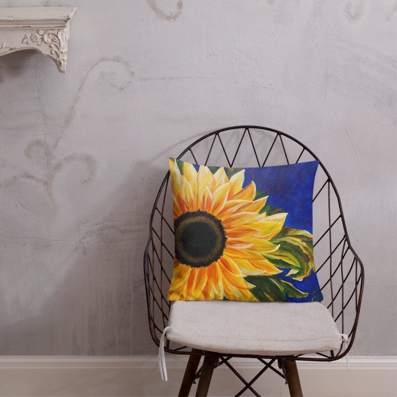 May include: A throw pillow with a sunflower design on a blue background. The sunflower is yellow with a dark brown center and green leaves. The pillow is sitting on a chair.