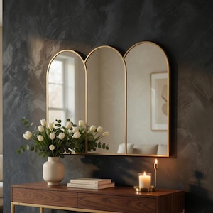 May include: A modern entryway scene featuring a dark wood console table with gold legs and trim. Above the table hangs a triple arched mirror with gold frames. A vase of white flowers and a lit candle add decorative touches.