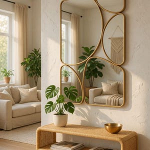 May include: A modern living room scene featuring a set of gold-framed mirrors in various shapes. A woven bench holds books and a gold bowl, with a potted plant in front. The room has neutral tones, with a sofa and plants visible.