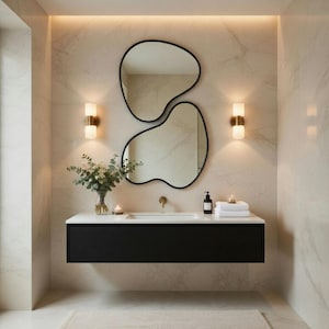 May include: A modern bathroom with a black floating vanity, a white countertop, and two organic-shaped mirrors with black frames. The walls are a light marble pattern, and there are two sconces with gold accents.