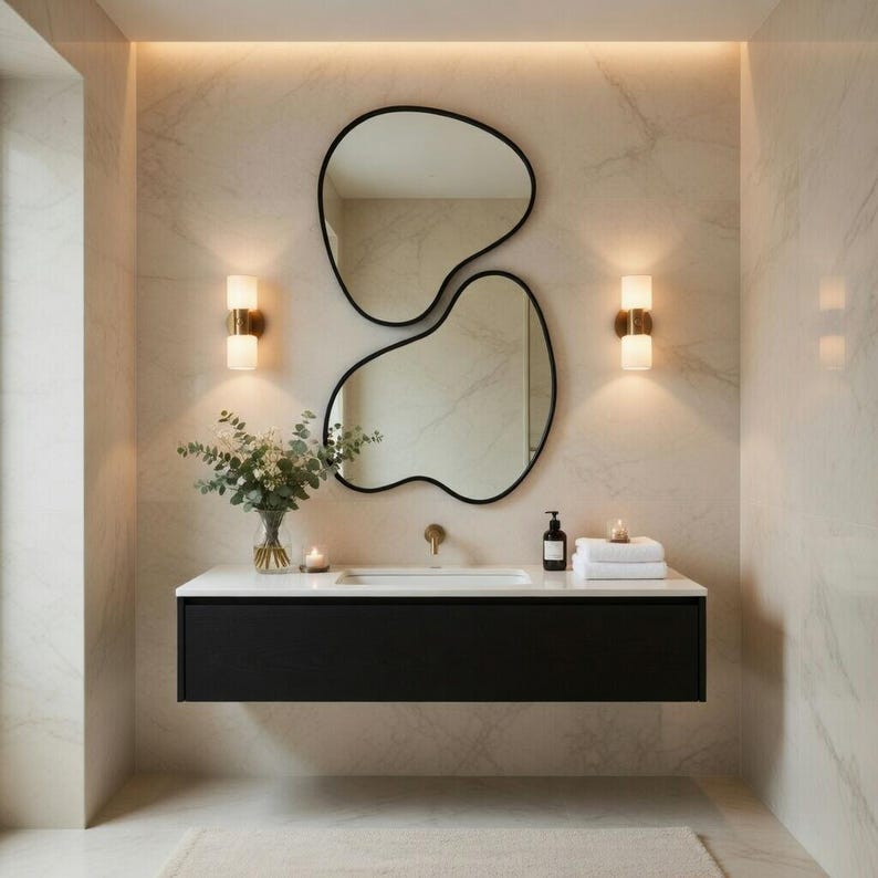 May include: A modern bathroom with a black floating vanity, a white countertop, and two organic-shaped mirrors with black frames. The walls are a light marble pattern, and there are two sconces with gold accents.