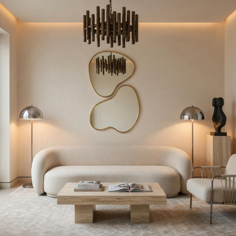 May include: A modern living room with neutral tones. A cream-colored sofa, a light-colored rug, and a travertine coffee table are the focal points. Two chrome floor lamps flank the sofa. A unique mirror and a dark bronze chandelier hang on the wall.