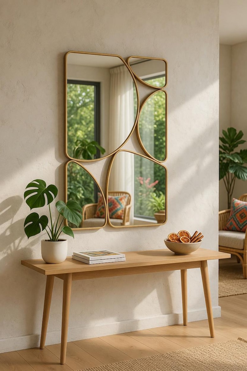 May include: A modern wall mirror with a gold frame, featuring an abstract design of connected rectangular and curved shapes. A wooden console table holds a potted plant, a book, and a bowl of dried fruit. The room has a neutral color palette.