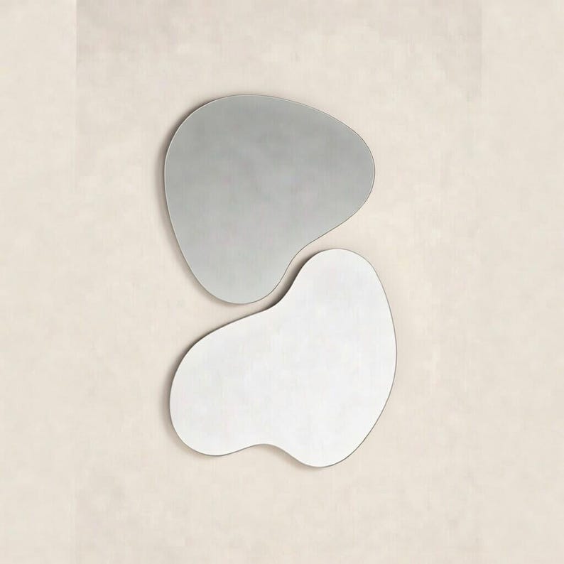 May include: Two wall mirrors with irregular, organic, amoeba-like shapes. The top mirror is slightly smaller with a greyish tint, while the bottom mirror is larger and brighter. Both pieces feature smooth, curved edges and a frameless design.
