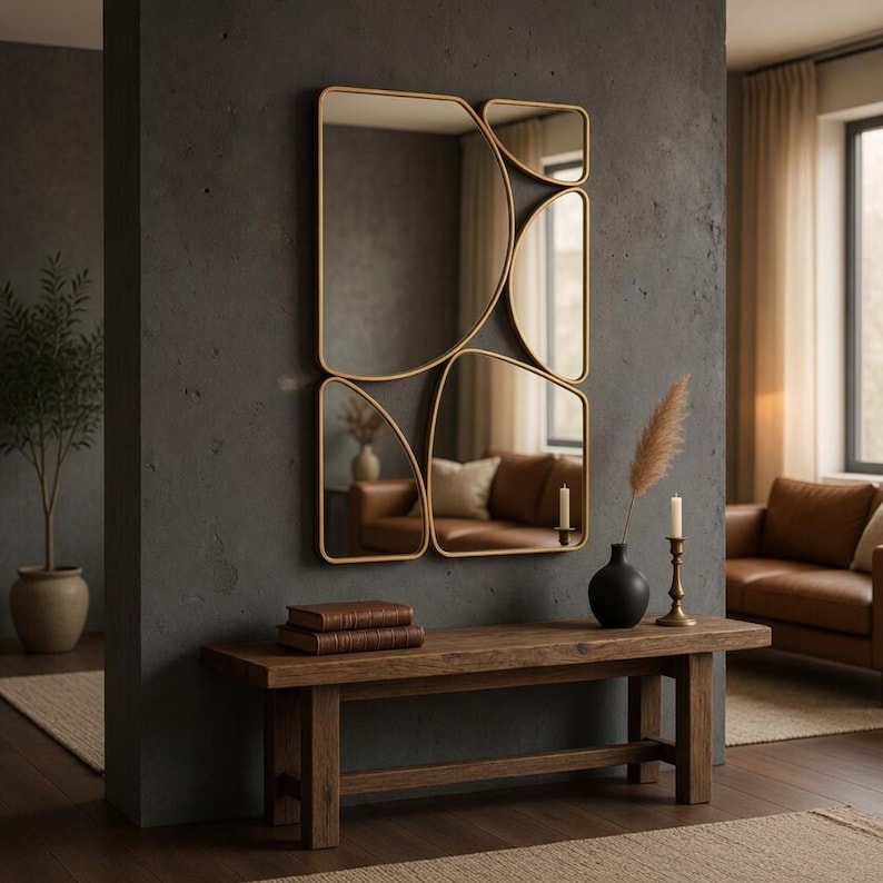 May include: A modern gold-framed mirror with an abstract design hangs on a dark gray wall. Below, a wooden bench holds books, a black vase with dried flowers, and candles. The room features a neutral color palette.