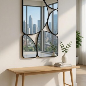 May include: A modern wall mirror with a unique design, featuring multiple irregularly shaped black-framed mirrors. The mirrors reflect a cityscape. Below, a light wood bench holds a white vase with greenery and books.