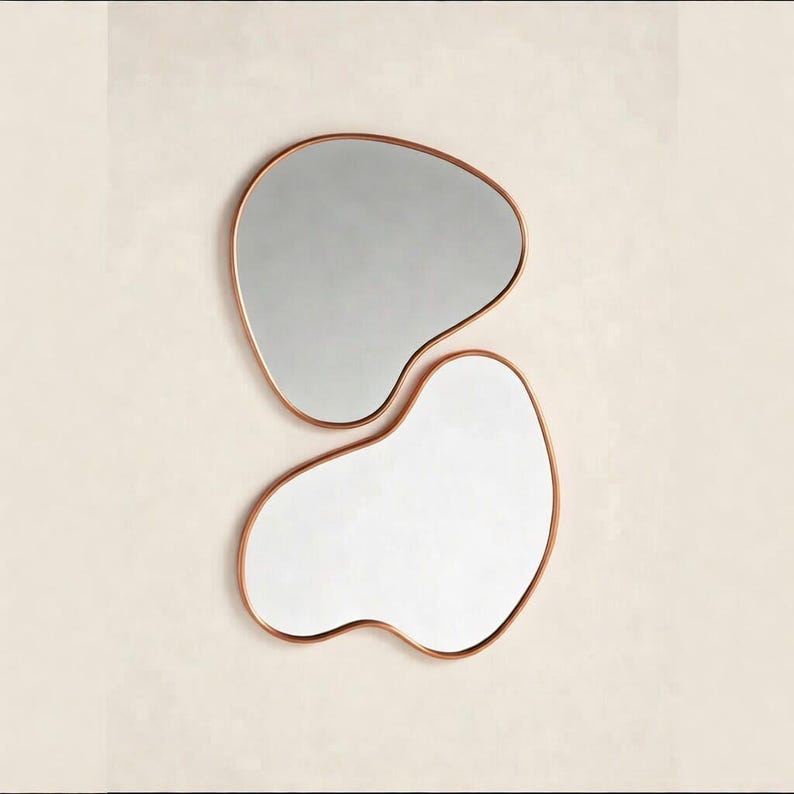May include: Two modern, abstract-shaped mirrors with copper-colored frames. The top mirror has a gray reflective surface, while the bottom mirror has a white reflective surface. The mirrors are mounted on a light beige wall.