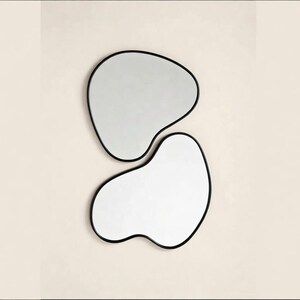 May include: Two modern, abstract mirrors with irregular, organic shapes. Each mirror has a reflective surface framed by a thin, black border. The mirrors are mounted on a neutral-colored wall, creating a minimalist aesthetic.