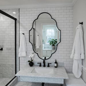 May include: A bathroom with a white marble countertop and a black faucet. A black-framed, ornate mirror hangs above the sink. White towels and a robe are present. The walls are white subway tile.