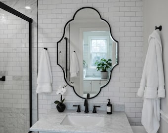 Puede incluir: Un baño con encimera de mármol blanco y grifo negro. Un espejo ornamentado con marco negro cuelga sobre el lavabo. Hay toallas y una bata blancas. Las paredes son de azulejos blancos.