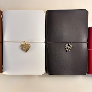 Puede incluir: Diarios de cuero en varios colores, incluyendo blanco y marrón oscuro. Cada diario está asegurado con un cordón y presenta un dije decorativo, como un corazón dorado o una hoja. Diseñados para escribir y tomar notas.