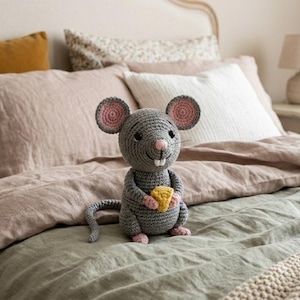May include: A gray crocheted mouse stuffed toy featuring large round ears with pink centers, a small pink nose, two white teeth, and a yellow triangular piece of cheese held in its front paws. The mouse has a long gray tail and small pink feet.