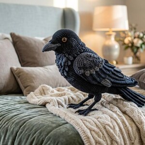Pode incluir: Uma figura de corvo feita à mão em crochê com fio azul-marinho escuro e preto, apresentando penas detalhadas nas asas, um peito texturizado e dois pequenos olhos castanhos. O pássaro é retratado em pé com a cabeça virada ligeiramente para a esquerda.