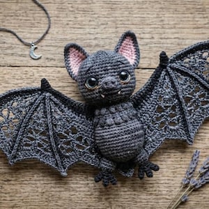 May include: A dark gray crocheted bat plush toy with large black eyes, small white fangs, and pink inner ears. The wings feature an intricate lace-like pattern. The bat is surrounded by two balls of gray yarn, a wooden crochet hook, and scissors.