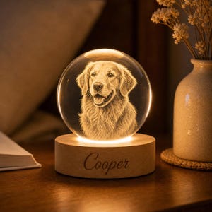 May include: A glowing crystal ball night light featuring a detailed engraving of a Golden Retriever dog. The light sits on a wooden base with the name "Cooper" engraved on it. The warm light illuminates the dog's image.