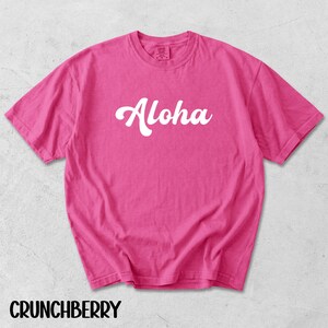 May include: A bright pink t-shirt with the word "Aloha" in white script across the front. The shirt has short sleeves and a crew neck. The word "CRUNCHBERRY" is printed in black at the bottom.