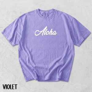 May include: Violet-colored t-shirt with the word "Aloha" in white script across the front. The shirt has short sleeves and a crew neck. The word "VIOLET" is printed in a sans-serif font at the bottom of the image.
