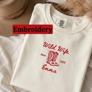 Könnte beinhalten: Cremefarbenes T-Shirt mit roter Stickerei. Der Text lautet "Wild Wife" über einem Paar Cowboy-Stiefel, mit "Juni" und "2026" sowie dem Namen "Emma" darunter. Das Wort "Stickerei" befindet sich in einem roten Rechteck.