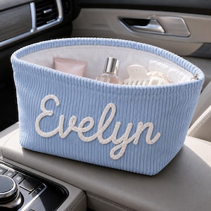 May include: A light blue corduroy storage basket with the name Evelyn embroidered in white script on the front. The interior is lined with white fabric and contains a small pink tube, a glass bottle, and a white hair clip for organization.