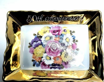 May include: A rectangular ceramic trinket dish with a scalloped edge and a metallic gold finish. The center features a white background with a colorful floral bouquet and the text 50th Anniversary written in a cursive white script font.