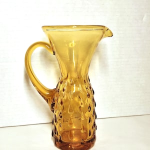 May include: An amber-coloured glass pitcher featuring a flared rim, a curved handle, and a textured exterior surface decorated with a repeating pattern of raised, teardrop-shaped indentations that wrap around the body of the vessel.