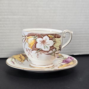 Puede incluir: Un juego de taza y platillo vintage. La taza blanca presenta un diseño floral con acentos amarillos, blancos y marrones. El platillo refleja el diseño de la taza, con ribete dorado en ambas piezas. El juego es probablemente de porcelana.