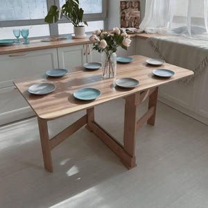 May include: A light-colored wooden dining table with a rectangular top and folding legs. The table is set with several plates in shades of blue and gray, and a vase of flowers. The table is approximately 48 inches long.