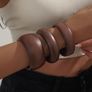 Könnte beinhalten: Drei dunkelbraune Holzarmreifen sind an einem Handgelenk gestapelt. Die Armreifen sind dick und haben eine glatte, polierte Oberfläche. Das Handgelenk ist sichtbar, zusammen mit einem Teil eines weißen Oberteils und einer schwarzen Jeans.