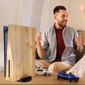 Op de afbeelding: Een houten console cover met een zwarte basis. De cover is gemaakt van lichtgekleurd hout met verticale nerf. Een persoon viert op de achtergrond, met twee gamecontrollers op de tafel.
