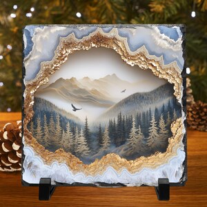 May include: Square stone art piece featuring a mountain landscape scene framed by a geode-like border. The artwork depicts a forest, mountains, and birds in flight, with a color palette of blues, browns, and golds. The piece is displayed on a black stand.