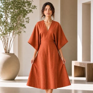May include: A rust-colored midi dress featuring a deep V-neckline, wide kimono-style sleeves, and a fitted waist that transitions into a flared A-line skirt. The garment is crafted from a solid-colored fabric with a subtle, natural-looking texture.