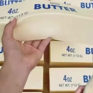 May include: A close-up of several 4 oz. salted butter packages. The packages are light yellow with blue text that reads "BUTTER" and "4 oz. NET WT. (113 G)". A hand is holding a butter package.