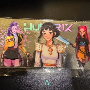 May include: A clear plastic package containing three anime-style character cutouts. The characters have various hair colours and outfits. The word "HURIX" is printed in a colourful font above the characters. The cutout on the left has purple hair and a yellow jacket.