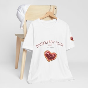 Puede incluir: Camiseta blanca con el texto "BREAKFAST CLUB PARIS EST. 1995" en borgoña. En la parte delantera, un pastel en forma de corazón con relleno rojo. Una gorra de béisbol blanca descansa sobre una silla de madera.