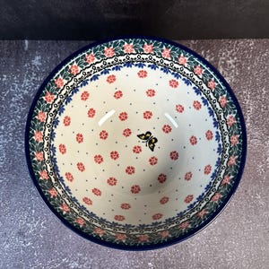 May include: A white ceramic bowl with a blue rim and floral patterns. The bowl features a repeating pattern of small red flowers and a black and yellow butterfly. The rim is decorated with pink flowers and green leaves.