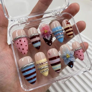 May include: A set of ten artificial nails in a clear plastic case. The nails feature various designs, including polka dots, stripes, and floral embellishments. Some nails have silver-colored bands and rhinestones.