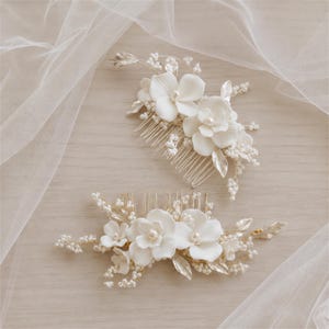 May include: Two white floral hair combs with pearl accents and silver-coloured leaves. The combs feature delicate, three-dimensional flowers, designed for bridal or special occasion hairstyles. The combs are displayed on a light-coloured wooden surface.