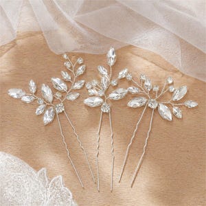 May include: Three silver hair pins with clear crystal accents. Each pin features a different floral design with various sizes of crystals. The pins are arranged on a light brown surface with a sheer white fabric in the background.