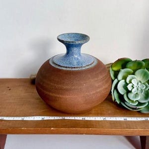 May include: A round ceramic vase with a matte brown body and a glossy blue speckled neck. A white measuring tape is positioned in front of the vase, showing a width of approximately 6 inches across the widest part of the ceramic vessel.