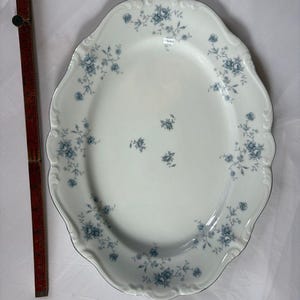 May include: An oval white porcelain serving platter with a scalloped edge and a blue floral pattern. A wooden folding ruler is placed vertically next to the platter, showing a length of approximately 14 inches along the side.