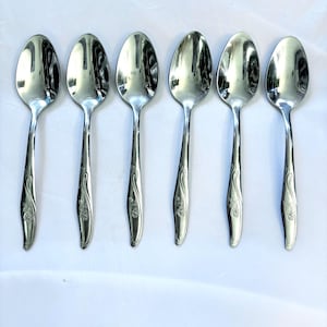 May include: A set of six matching silver-toned metal spoons arranged in a row. Each spoon features a smooth, oval-shaped bowl and a slender handle decorated with a subtle, embossed floral pattern near the base of the stem.