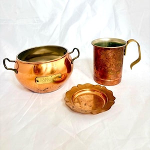 May include: A copper potpourri bowl with two brass handles and a brass label reading POTPOURRI, a copper mug with a brass handle, and a small round copper tray with a scalloped edge. All items feature a metallic finish.