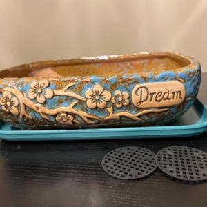 May include: A ceramic planter with a blue and brown glaze, featuring embossed floral designs and the word "Dream." The planter sits on a matching blue tray, with two black mesh inserts nearby. A decorative item for plants.