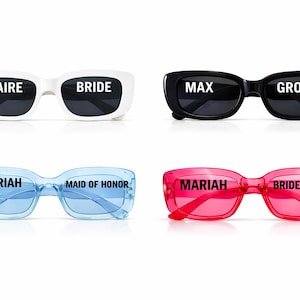 May include: Four pairs of sunglasses in white, black, blue, and pink. Each pair has text on the lenses: "CLAIRE BRIDE", "MAX GROOM", "MARIAH MAID OF HONOR", and "MARIAH BRIDESMAID". The sunglasses are a fun accessory for a wedding party.
